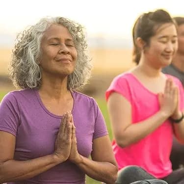 People sitting outdoors with their eyes closed and hands together.