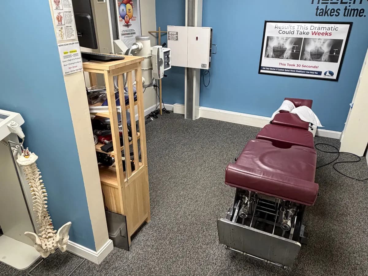 An adjusting room at Active Chiropractic in Saco: a burgundy chiropractic table on grey carpet in front of a blue wall that reads "Healing takes time", with a spine model beside it