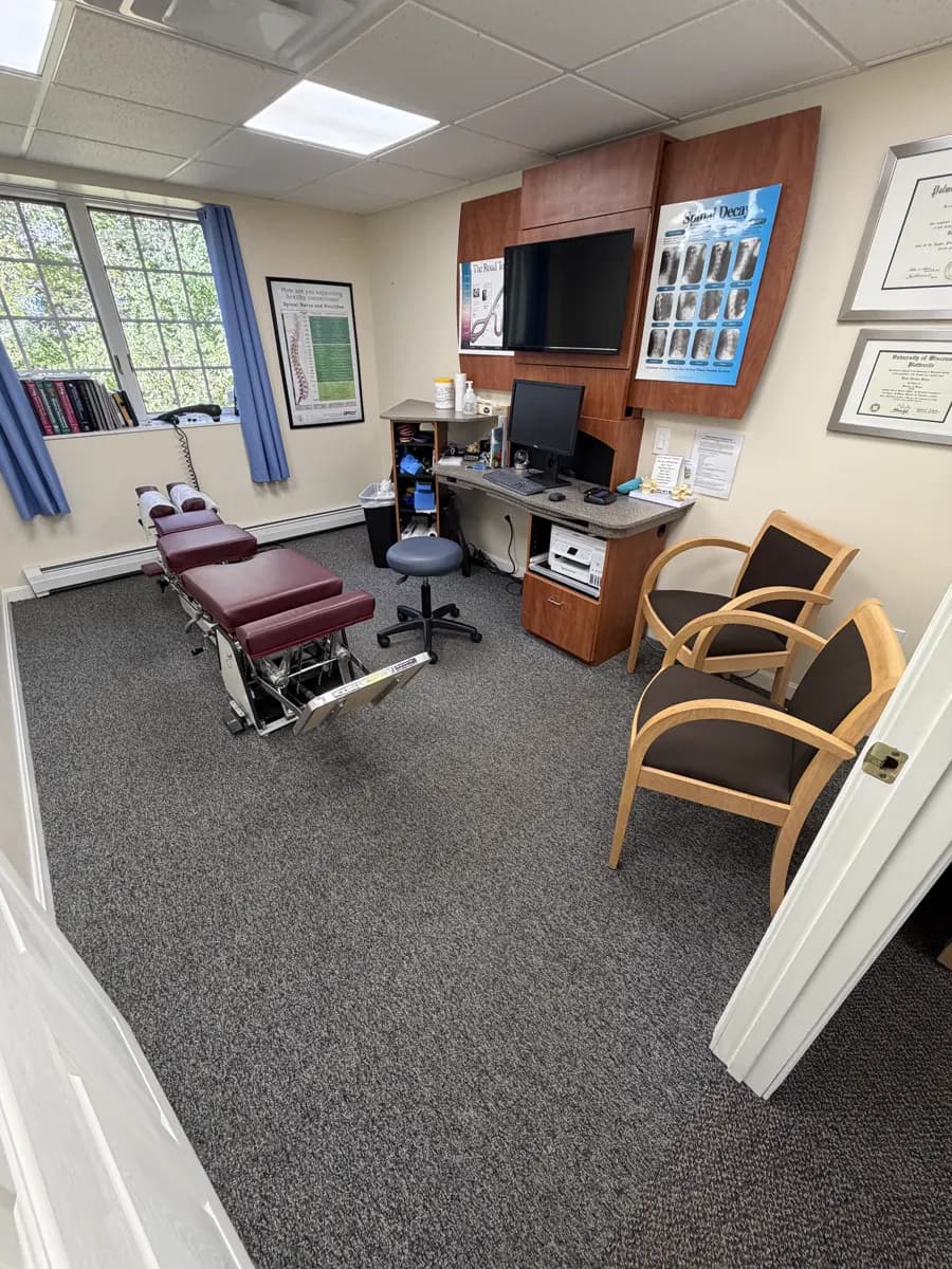 The consultation room: an adjusting table, a desk with a computer and a wall screen, two chairs and framed diplomas.