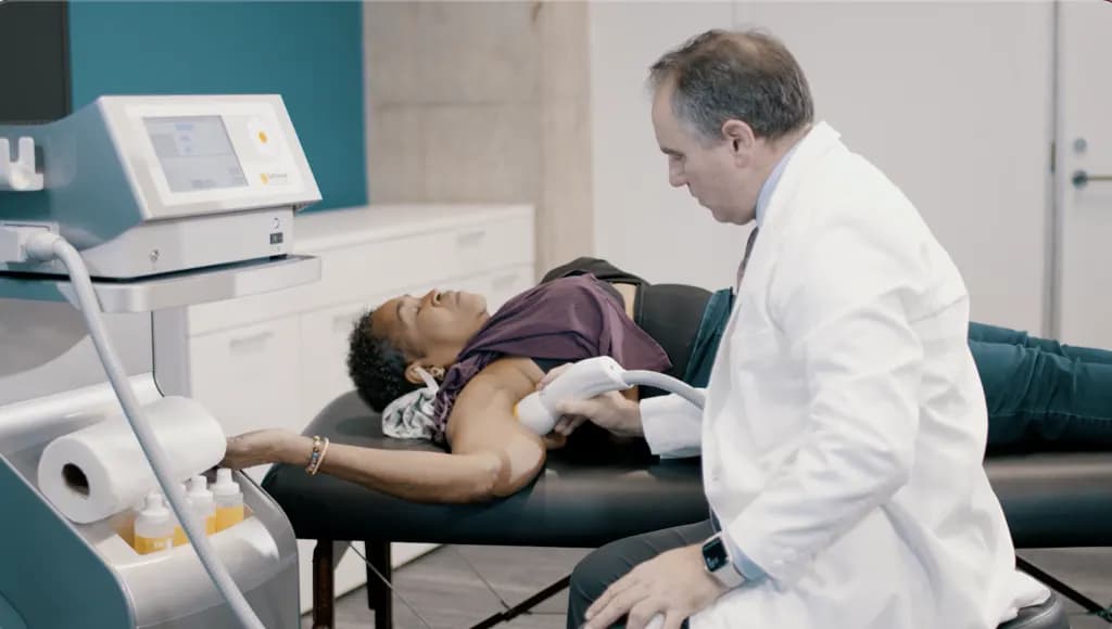 A practitioner holding the SoftWave applicator to the shoulder of a patient lying on a treatment table, with the SoftWave unit beside them